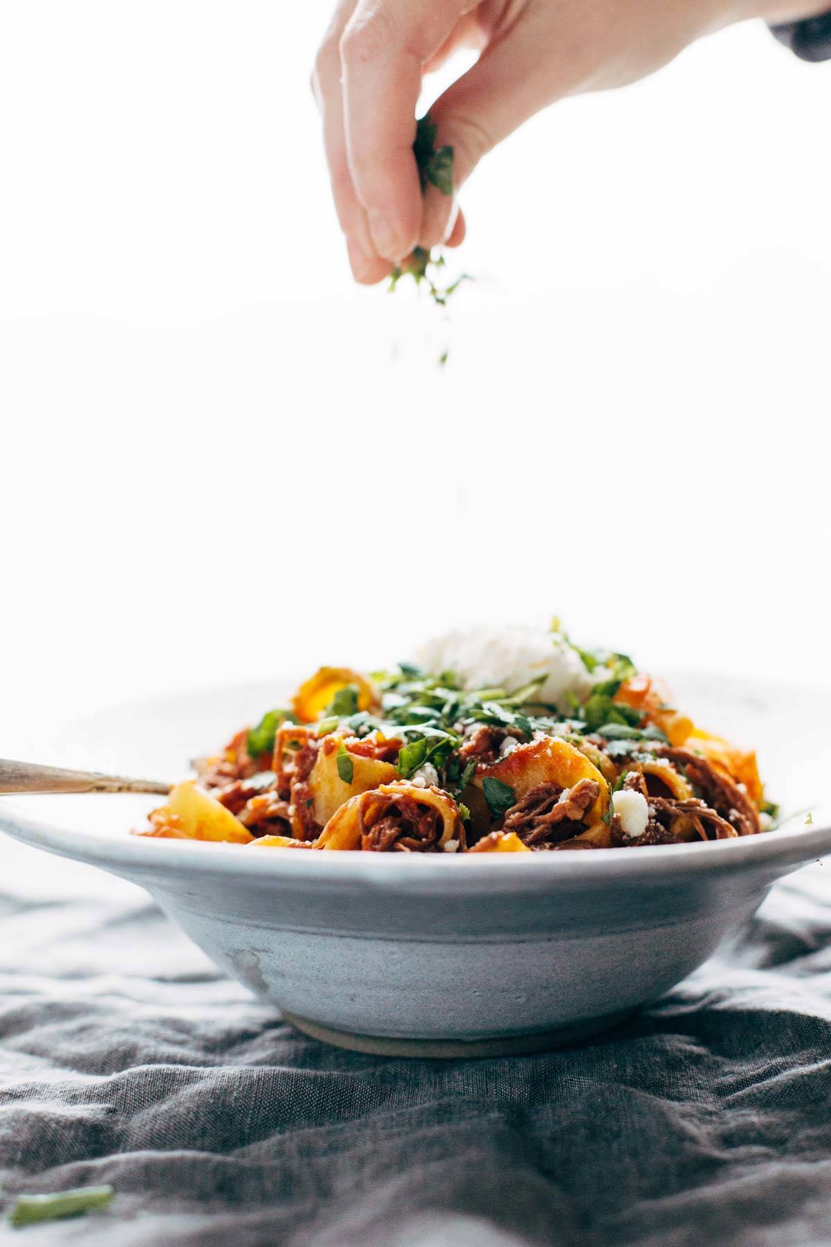 A side angle view of beef ragu in a bowl with a fork. A white hand is sprinkling herbs on top.