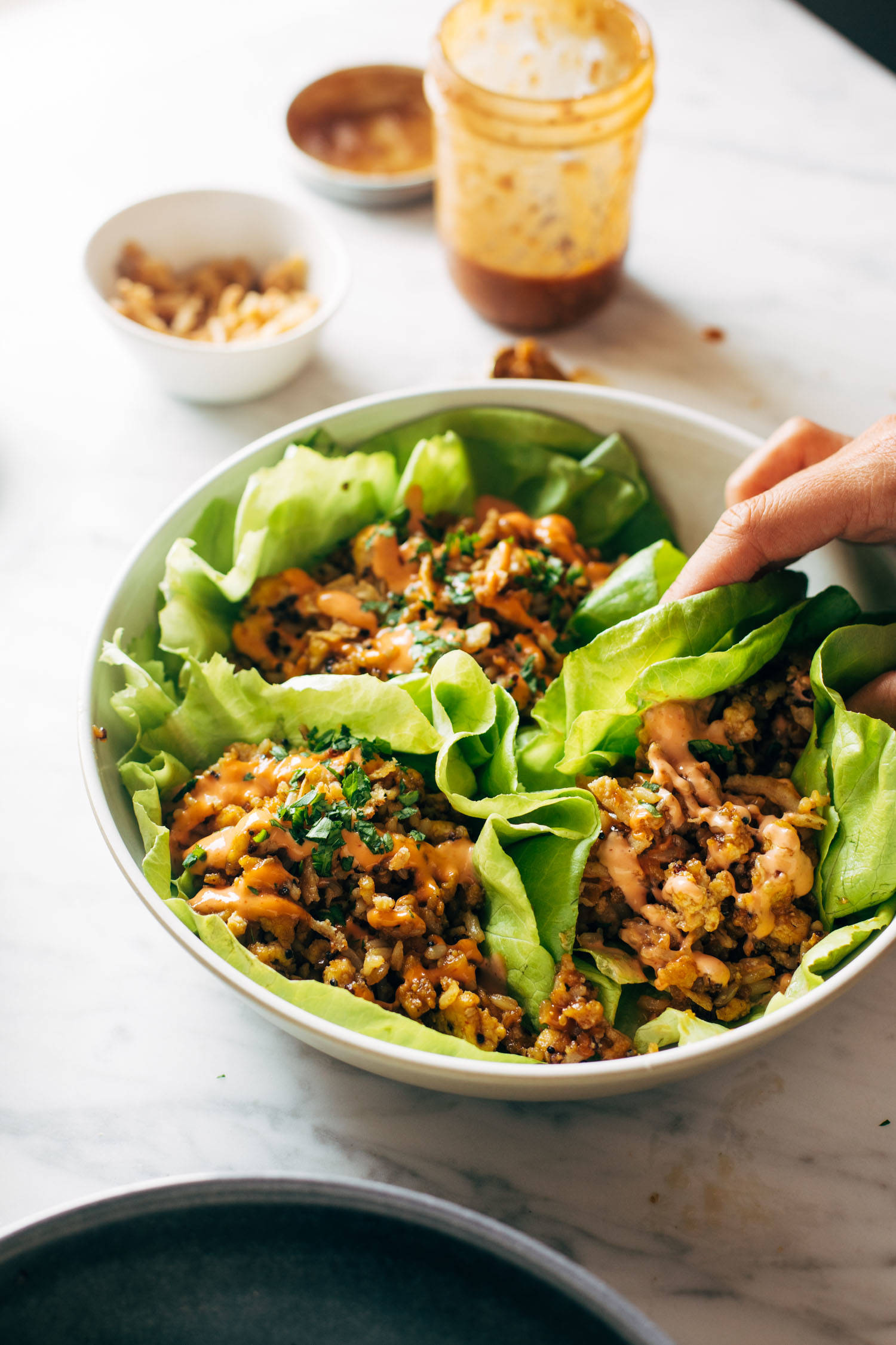 A white hand grabbing a tofu and brown rice lettuce wrap on a platter. There are extra ingredients for the lettuce wraps in the background.